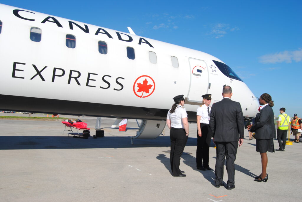 Group of people near an Air Canada Express airplane on the tarmac, with some in uniform and others in business attire, under a clear blue sky.