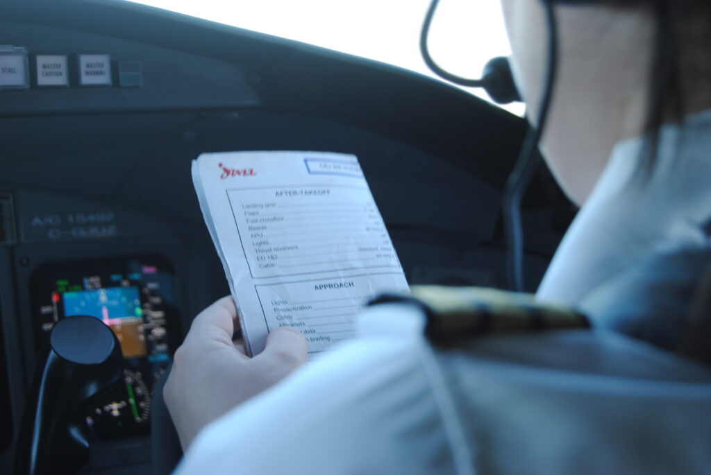 Pilot in uniform holding an 'AFTER TAKEOFF' checklist inside an aircraft cockpit, with flight instruments and screens visible.