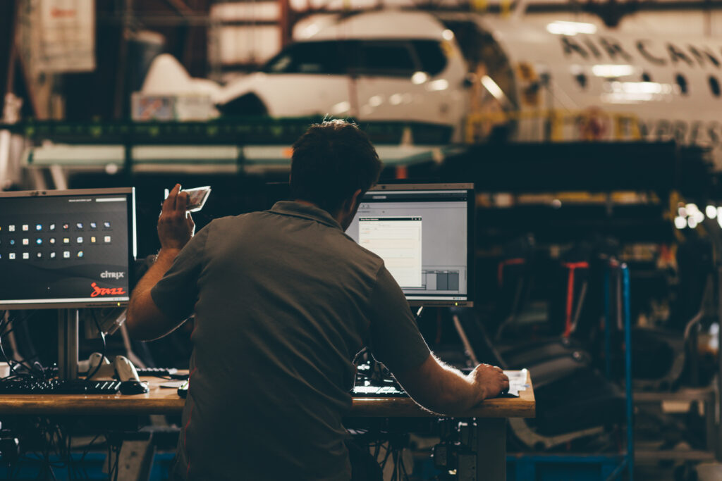 Person working at a desk with two monitors in an aircraft maintenance facility, with tools, equipment, and a partially visible airplane fuselage in the background.