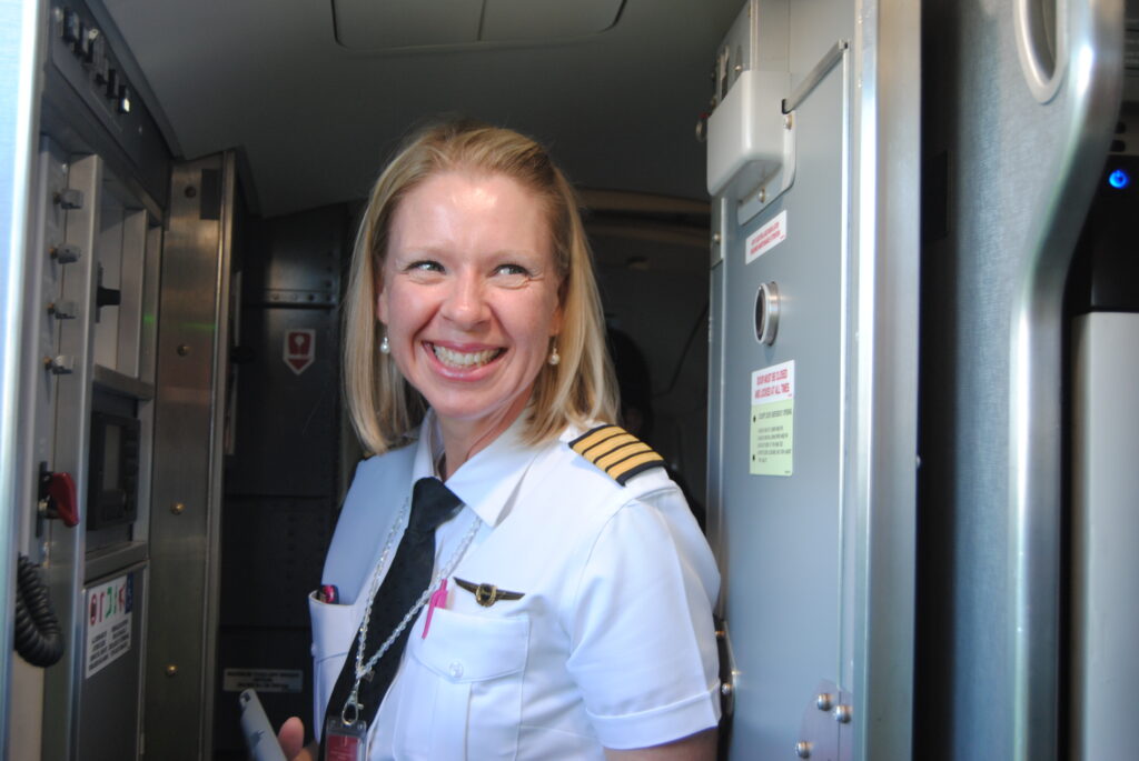Woman in pilot uniform standing on aircraft, smiling.