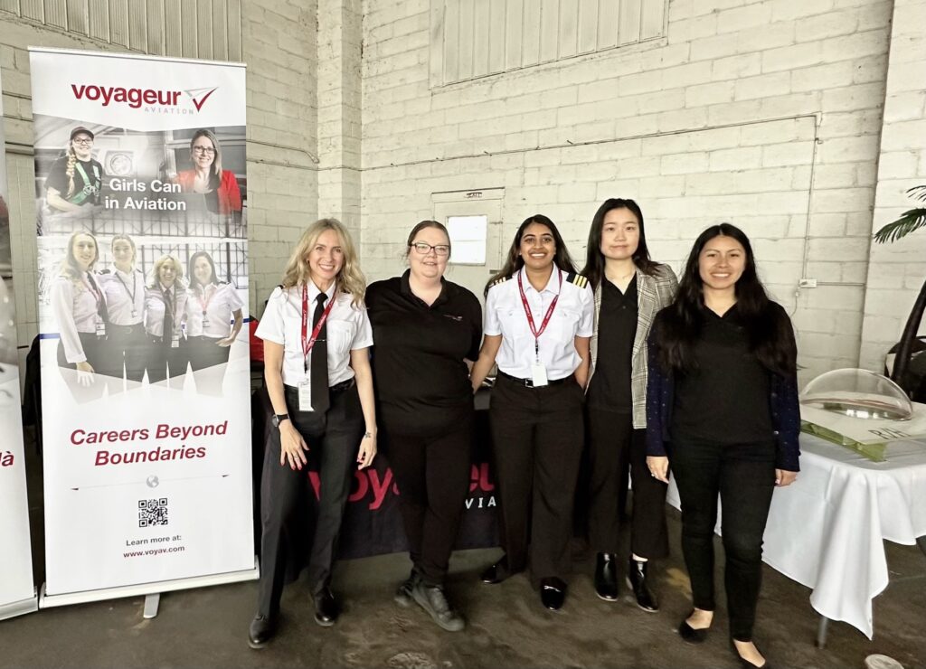Five people standing in front of a 'Girls Can in Aviation' banner by Voyageur Aviation, promoting careers in aviation; a table and QR code are also visible.